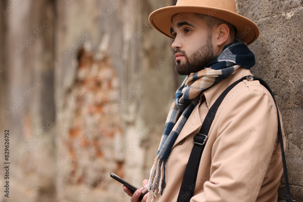 Obraz premium Handsome man in warm scarf with smartphone near wall outdoors. Space for text