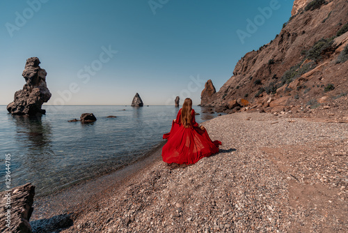 A woman in a red dress stands on a beach with a rocky shoreline in the background. The scene is serene and peaceful  with the woman s red dress contrasting against the natural elements of the beach.