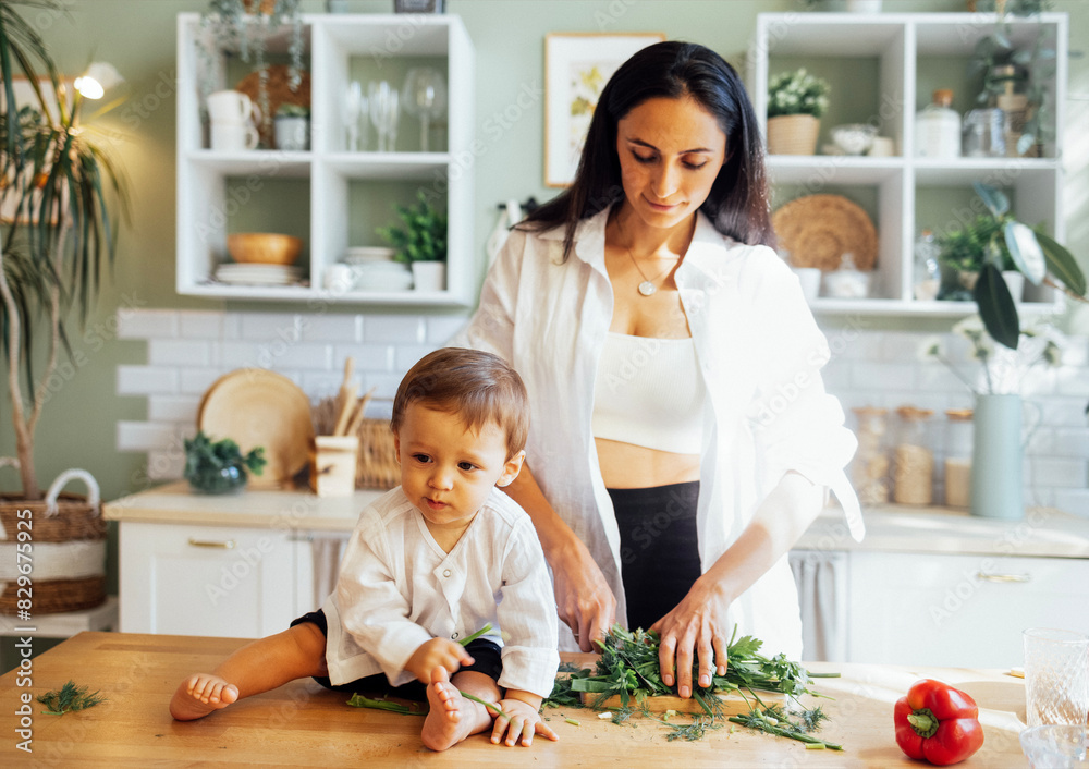 Fototapeta premium A young mother is cutting greens on a wooden stand in a modern kitchen and her little baby is playing next to her on the table.