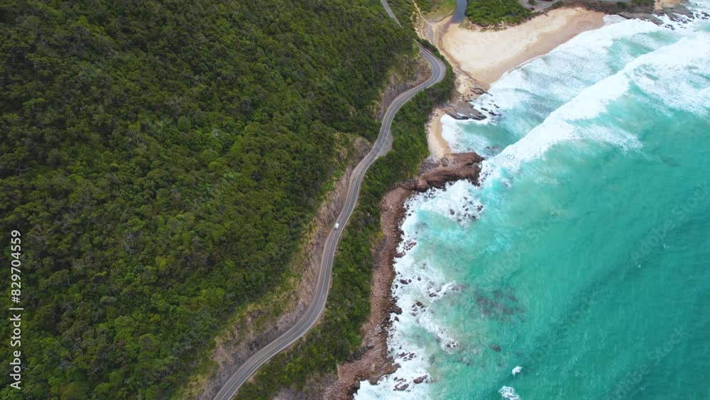 4K Drone view of a camper van driving along the Great Ocean Road in Victoria, Australia. The drone pans up to reveal a stunning Australian landscape. The blue ocean in view next to the green cliffs.