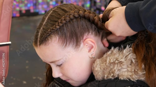 A young girl sits quietly as her hair is intricately braided in a salon. She wears a black, fur-lined jacket photo