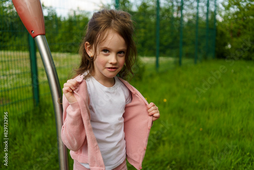 A little smiling, beautiful girl takes off a sweater on a summer playground.