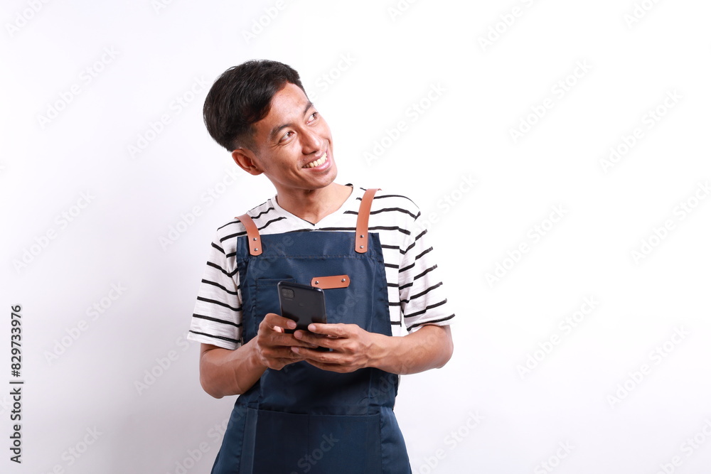 Young asian man barista bartender barman employee in blue apron hold mobile cell look aside isolated on white background Young asian man barista bartender barman employee in blue apron hold mobile cell look aside isolated on white background
