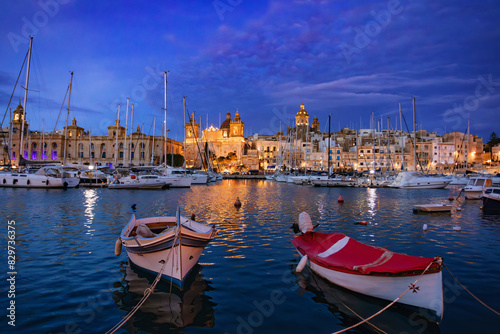 Senglea city, part of the Three Cities of Malta. Night marina glows under the night sky, yacht masts silhouetted against twinkling lights reflecting on the tranquil waters. Travel destination.