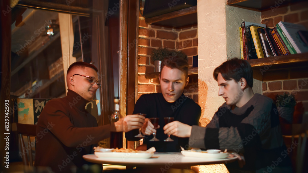 Three men are drinking in restaurant. Media. Friends are sitting in cozy bar and drinking alcoholic drink. Friends at bar clink glasses and drink