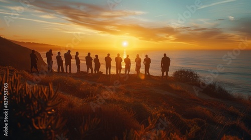 A group of people are standing on a hill overlooking the ocean. The sun is setting, casting a warm glow over the scene. The people are all looking out at the water, taking in the beauty of the moment