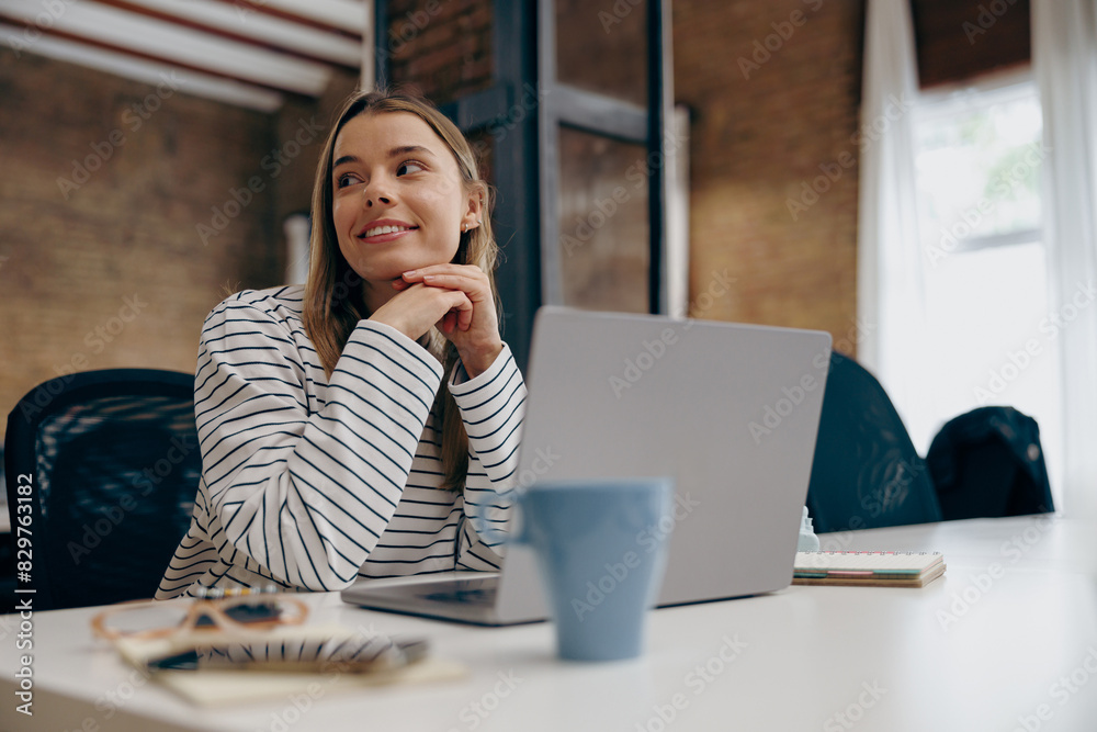 Pretty young woman manager working on laptop while sitting the desk in office and looks away Pretty young woman manager working on laptop while sitting the desk in office and looks away