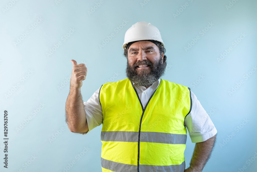 smiling adult man engineer with beard and wearing helmet and safety vest looking at camera on blue background, using his tablet and reviewing work, pointing at something or someone smiling adult man engineer with beard and wearing helmet and safety vest looking at camera on blue background, using his tablet and reviewing work, pointing at something or someone