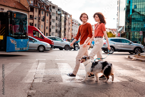 Side view of young couple, man and woman, are walking through city through pedestrian crossing with their dog. Sunset light in background