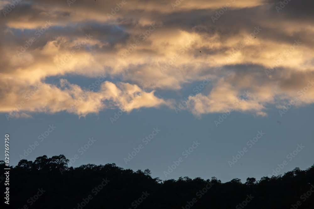 Landscape of thick clouds, orange light hitting the blue sky, silhouettes of trees, forests, and mountains. Area for text in the middle of the image Landscape of thick clouds, orange light hitting the blue sky, silhouettes of trees, forests, and mountains. Area for text in the middle of the image