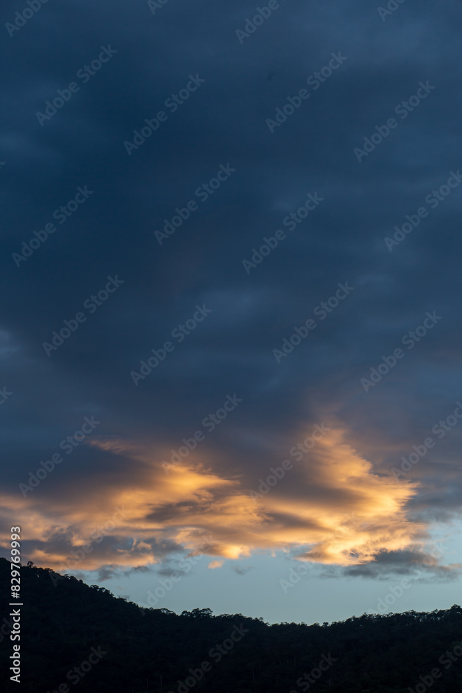 Landscape of thick clouds, orange light hitting the blue sky, silhouettes of trees, forests, and mountains. Area for text in the middle of the image