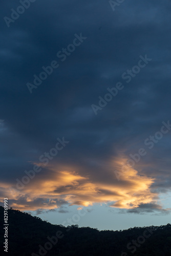 Landscape of thick clouds  orange light hitting the blue sky  silhouettes of trees  forests  and mountains. Area for text in the middle of the image