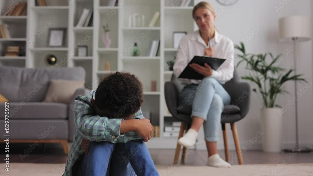 Disappointed little child in the psychologist office. Portrait of little boy sitting on floor with his back to the psychologist with his legs crossed during therapy session. Concept of child autism. Disappointed little child in the psychologist office. Portrait of little boy sitting on floor with his back to the psychologist with his legs crossed during therapy session. Concept of child autism.