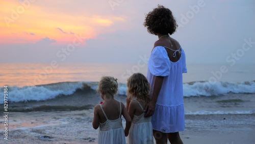 Mother and twins stand on shore endless sea, admiring sunset. Vibrant colors sunset reflected in water, creating magnificent spectacle. Mother hugs children watch sun slowly disappear below horizon.