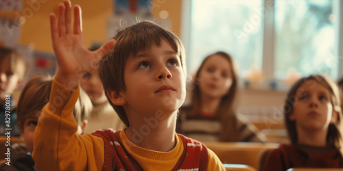 A boy in a yellow sweater raises his hand in a classroom. The other children are watching him