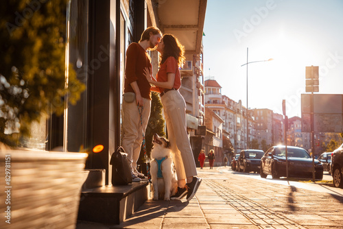 Young couple Caucasian man and woman, are kissing on street. Dog is sitting next to them on leash. Sunset light city in background