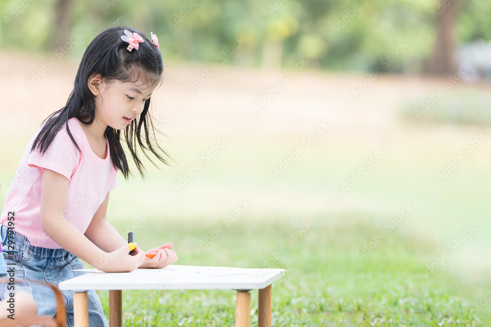 Happy Asian child girl coloring with crayons on paper at park, sitting on grass. Little cute kid with smiling face have fun outdoors activities. Creatives children. Education Happy Asian child girl coloring with crayons on paper at park, sitting on grass. Little cute kid with smiling face have fun outdoors activities. Creatives children. Education