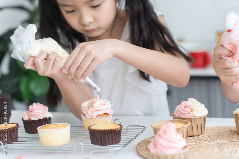 Asian little cute child girl enjoy making cupcakes with friend at home kitchen, squeezing cream from piping bag to decorate homemade cupcake, smiling with holiday activities. Selective focus on hands