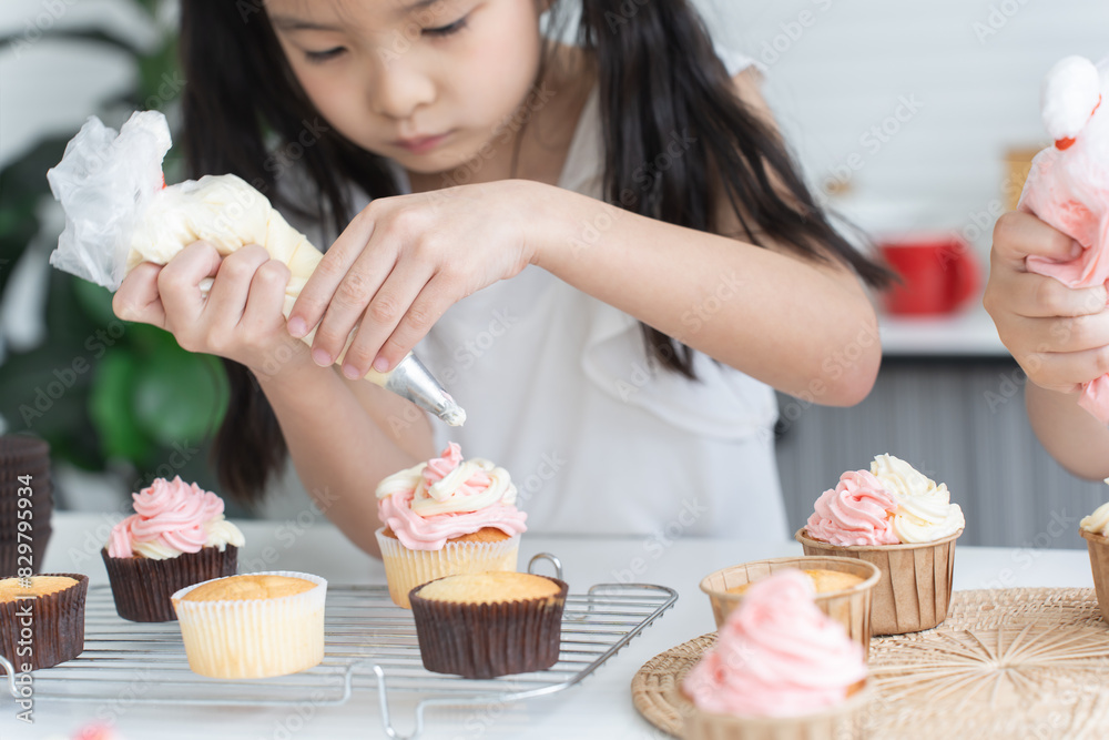 Asian little cute child girl enjoy making cupcakes with friend at home kitchen, squeezing cream from piping bag to decorate homemade cupcake, smiling with holiday activities. Selective focus on hands Asian little cute child girl enjoy making cupcakes with friend at home kitchen, squeezing cream from piping bag to decorate homemade cupcake, smiling with holiday activities. Selective focus on hands