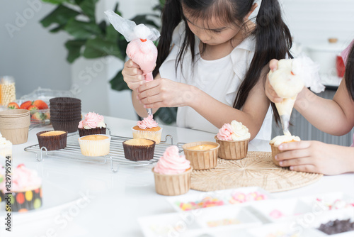 Billede på lærred Asian little cute child girl enjoy making cupcakes with friend at home kitchen, squeezing cream from piping bag to decorate homemade cupcake, smiling with holiday activities