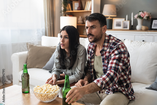 Excited and tense young couple sitting on sofa at home watching or streaming sports event on tv together. Man and woman watch football or soccer game on television, feeling nervous and waiting.