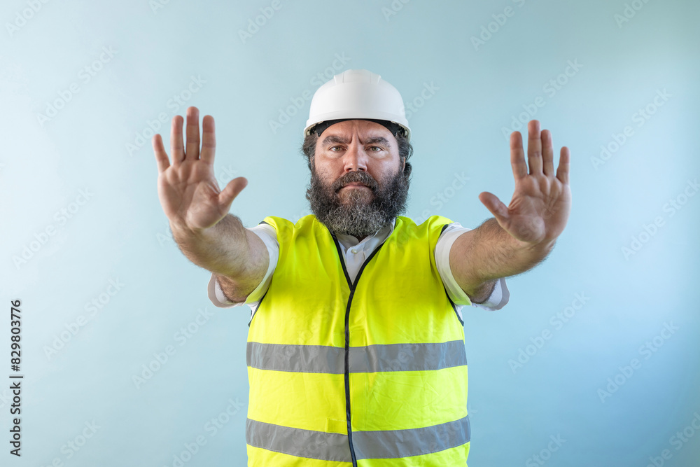 serious adult man engineer with beard and wearing helmet and safety vest looking at camera on blue background, making the stop sign with your hands serious adult man engineer with beard and wearing helmet and safety vest looking at camera on blue background, making the stop sign with your hands