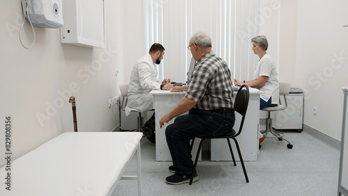 Senior man patient sitting on chair by the table of a doctor and a nurse. Clip. Medical checkup visit at the hospital.