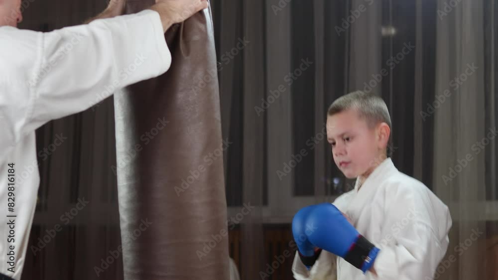 A boy in blue arm pads throws punches on a large hanging machine with the help of a trainer A boy in blue arm pads throws punches on a large hanging machine with the help of a trainer