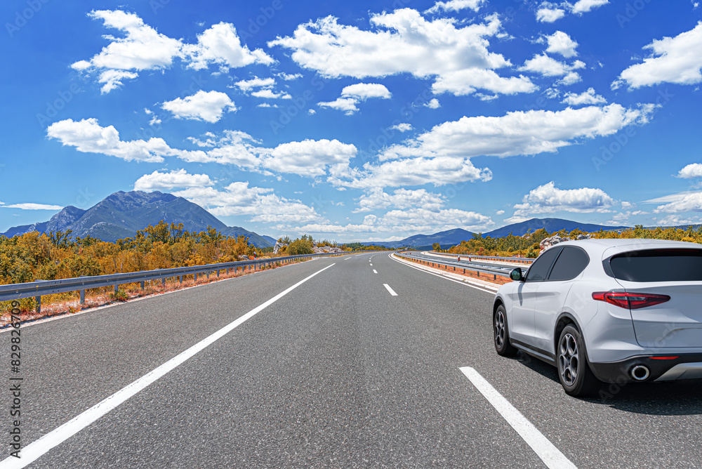 A white car drives along the highway against the backdrop of rocky mountains on a sunny day.