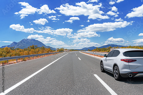 Obraz na płótnie A white car drives along the highway against the backdrop of rocky mountains on a sunny day. w salonie