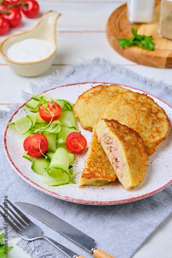 Potato pancakes stuffed with minced meat on a ceramic plate on a light wooden background. Served with fresh cucumber and cherry tomatoes. Belarusian cuisine. Potato pancakes stuffed with minced meat on a ceramic plate on a light wooden background. Served with fresh cucumber and cherry tomatoes. Belarusian cuisine.