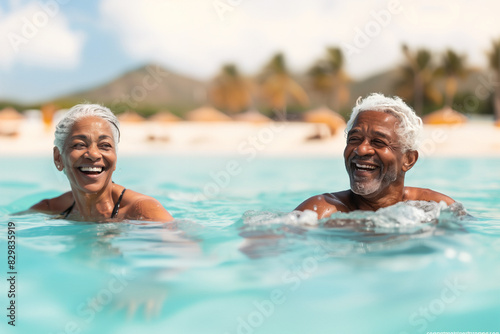 An elderly couple with gray hair enjoys a refreshing swim in clear blue waters at a tropical beach. The background features palm trees and distant hills under a bright, sunny sky. © SnapVault
