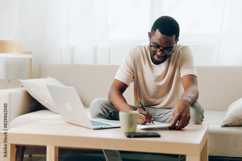 Smiling African American Freelancer Working on Laptop in a Modern Home Office, Typing and Enjoying the Cyberspace Lifestyle This image depicts an African American man sitting on a comfortable sofa in Smiling African American Freelancer Working on Laptop in a Modern Home Office, Typing and Enjoying the Cyberspace Lifestyle This image depicts an African American man sitting on a comfortable sofa in