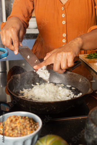 Close-up of Hispanic woman's hand in kitchen pouring chopped onion into frying pan with oil