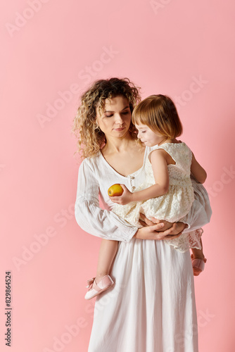 Woman holding child in white dress on pink background.