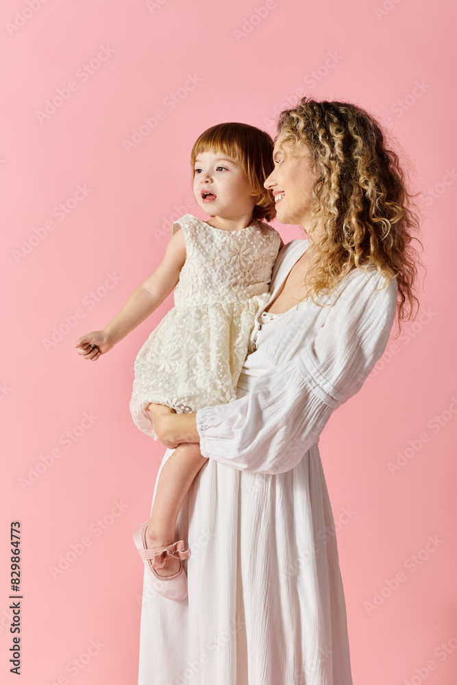 A mother in a white dress lovingly holds her baby girl in a white dress on a soft pink background. A mother in a white dress lovingly holds her baby girl in a white dress on a soft pink background.