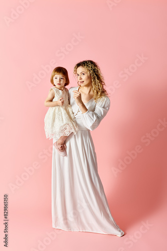 A woman in white dress holds a baby girl in a white dress against a pink background.