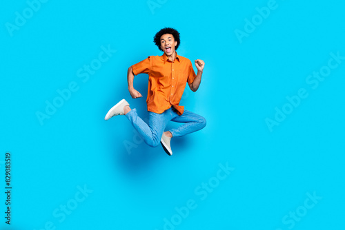 Full length photo of funky cool guy dressed orange shirt jumping high emtpy space isolated blue color background