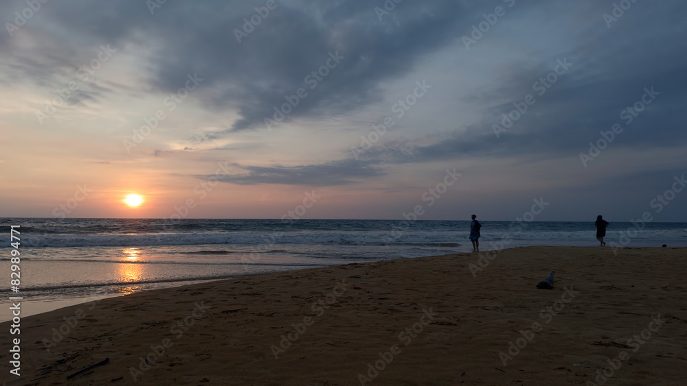 People are standing on sand with view of sea sunset. Action. People look at sunset from sandy shore. Beautiful landscape with people on sand on background of waves and setting sun