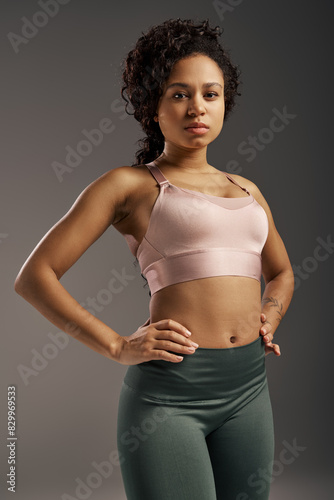 Curly African American sportswoman in pink sports bra and green leggings working out in studio with grey background. © LIGHTFIELD STUDIOS