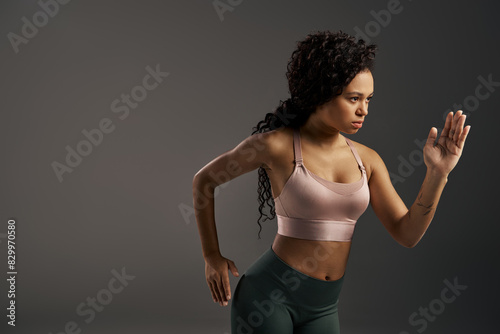 Curly African American sportswoman in sports bra and leggings striking a pose in a photo shoot with a grey backdrop.