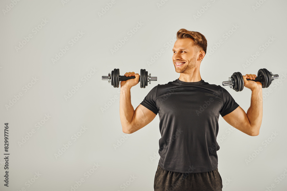 A young sportsman in active wear holds two dumbbells in front of a gray background in a fitness studio.