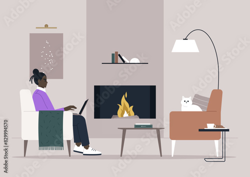 A person enjoys working on their laptop while sitting in a chair by the warm fireplace, with a cat resting nearby on another chair, and a book on the coffee table
