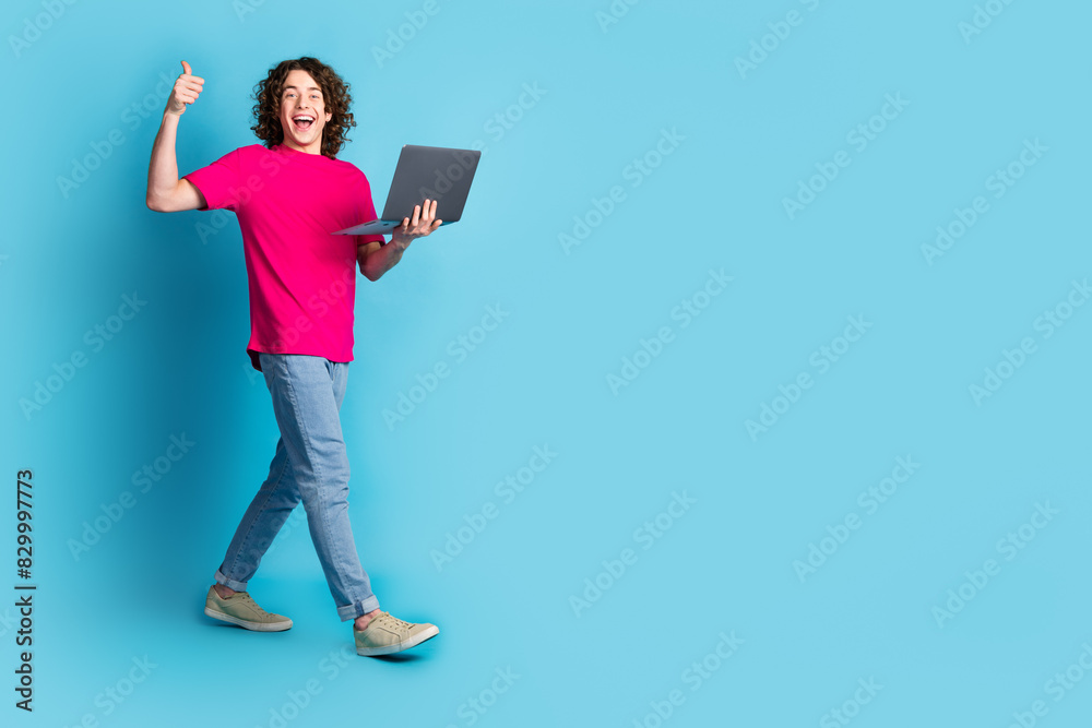 Full length portrait of nice young man walk netbook thumb up empty space wear t-shirt isolated on blue color background