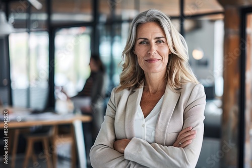 A portrait of a mature, successful senior woman boss inside the office in a business suit shows a businesswoman smiling and looking at the camera, standing with crossed arms.