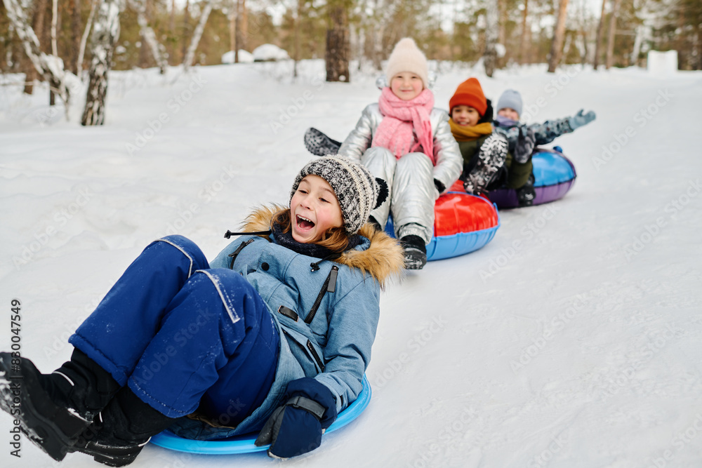 Cute excited boy in winterwear sitting on slide while moving down snow hill against group of his cheerful friends on tubes Cute excited boy in winterwear sitting on slide while moving down snow hill against group of his cheerful friends on tubes