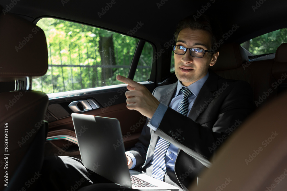 Successful handsome businessman sitting in the passenger seat in his luxury car working on laptop and driving to a business meeting