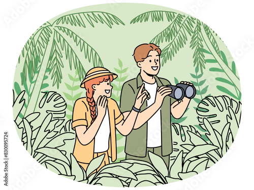 Tourists participate in jungle safari standing among dense tropical vegetation and holding binocular in hands. Happy couple of man and woman went on safari journey to explore wildlife