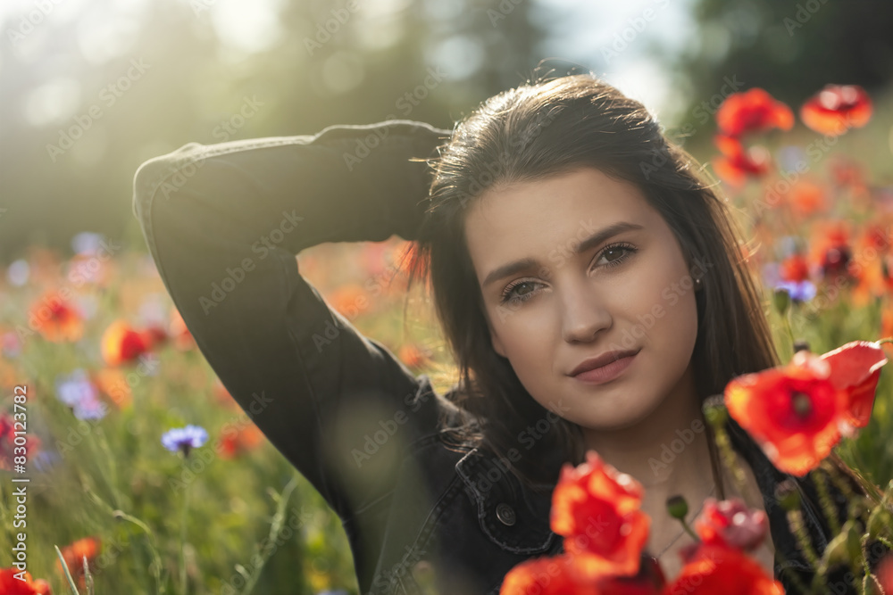 Naklejka premium Portrait of sadly smiling brunette girl with her hand in hair in red poppies field. Horizontally. 