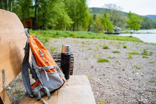 A backpack is resting on a wooden bench in a picturesque landscape, next to a water bottle. The grassy ground meets the asphalt road under the clear blue sky, surrounded by trees © Aleksey